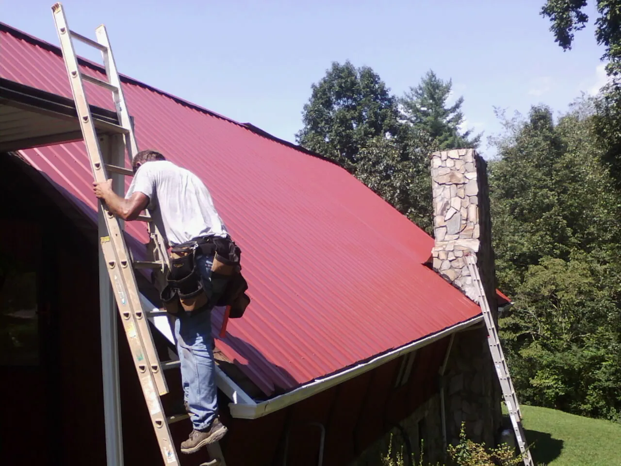 Roofer on a ladder beside a red metal roof and stone chimney — Soffit & Fascia Repair by Roofing Brothers in Mc Kee